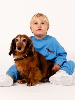 Front view of a child wearing a blue sweatshirt with a dog embroidery on the chest and matching blue trousers sitting next to a dog.