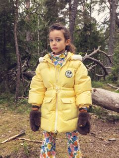 Yellow heavy puffer jacket with a Ritzratz patch, worn in a forest with brown furry mittens.