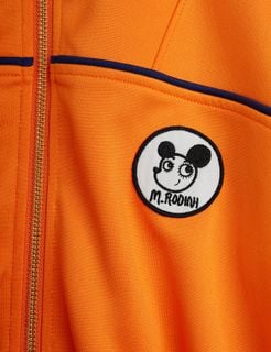 Orange recycled polyester tracksuit jacket detail featuring a circular Ritzratz patch and gold zipper.