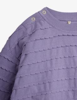 Close-up of a purple bodysuit featuring textured scalloped frill details and gold snap buttons at the shoulder.