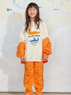 Orange recycled polyester trousers with front creases, Ritzratz patch, and striped side-piping, worn by a child.