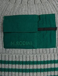 Close-up of a green woven pocket with logo embroidery on a grey ribbed hat with green stripes.