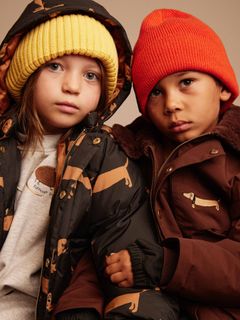 Two children wearing black and brown puffer jackets featuring a Dog print and colorful beanies.