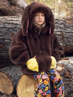 Brown faux fur jacket with a rabbit-ear hood and yellow pockets, worn by a child sitting on a log.