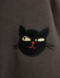 Close-up of a grey sweatshirt featuring an embroidered black cat motif with yellow eyes and orange nose.