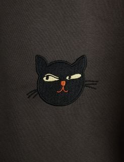 Close-up of a grey sweatpant fabric featuring an embroidered black cat face motif with yellowish eyes and an orange nose.