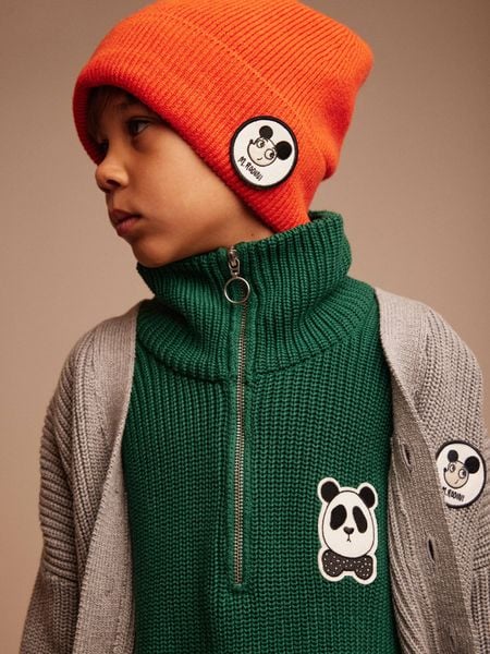 Child wearing a red beanie with a Ritzratz patch and a green zip-up cardigan.