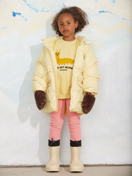 Yellow sweatshirt featuring a Velvet worm print, worn with a yellow puffer jacket and brown mittens.
