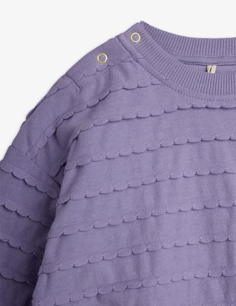 Close-up of a purple bodysuit featuring textured scalloped frill details and gold snap buttons at the shoulder.