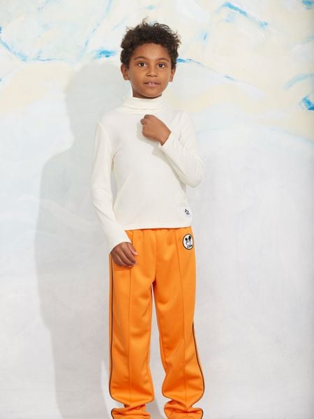 Orange Ritzratz patch trousers in recycled polyester with side piping, worn by a child with a cream turtleneck.