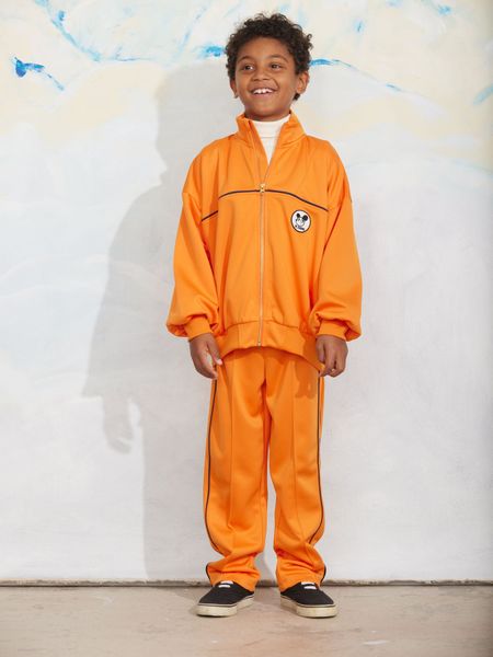 Orange recycled polyester trousers with a Ritzratz patch, striped side-piping and front creases, worn by a child.