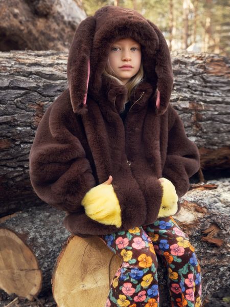 Brown faux fur jacket with a rabbit-ear hood and yellow pockets, worn by a child sitting on a log.