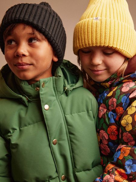 Two children wearing green and patterned puffer jackets and beanies.