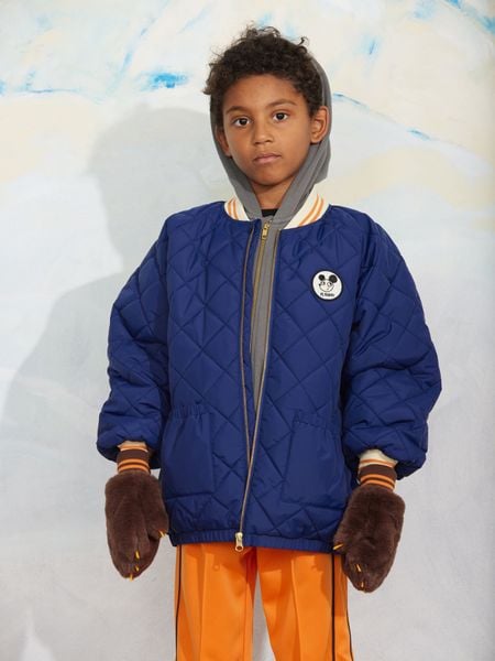 Blue quilted puffer jacket with a Ritzratz patch, zipper closure, and front pockets, worn with furry brown mittens.