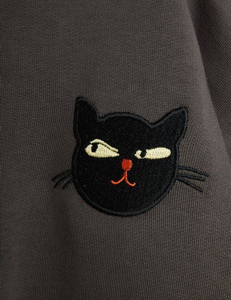 Close-up of a grey sweatshirt featuring an embroidered black cat motif with yellow eyes and orange nose.