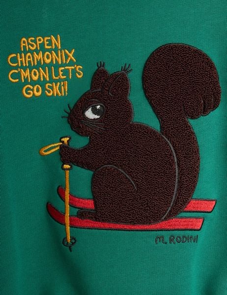 Close-up of a green sweatshirt featuring a brown chenille skiing squirrel patch with skis and yellow text.